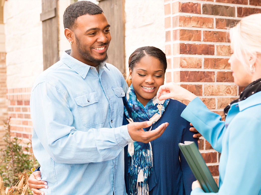 A young Black male and female couple standing in front of a home being handed keys by another woman.