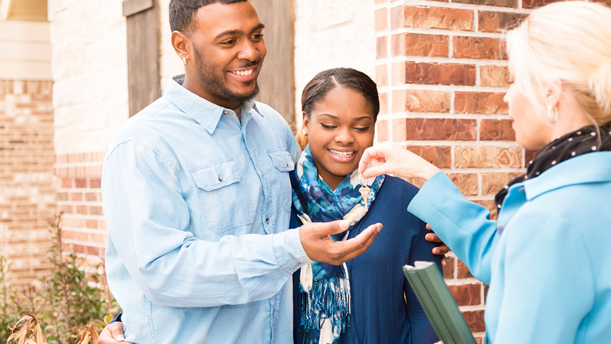 A young Black male and female couple standing in front of a home being handed keys by another woman.