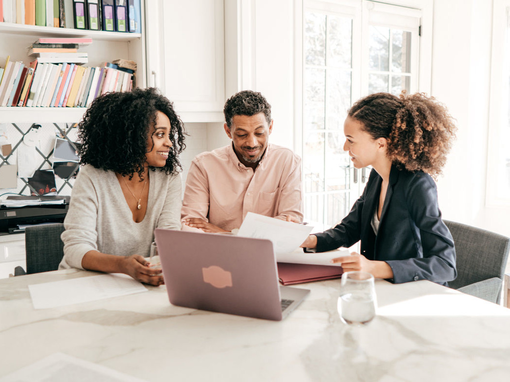 A male and female couple sit at a table with another woman showing them documents.