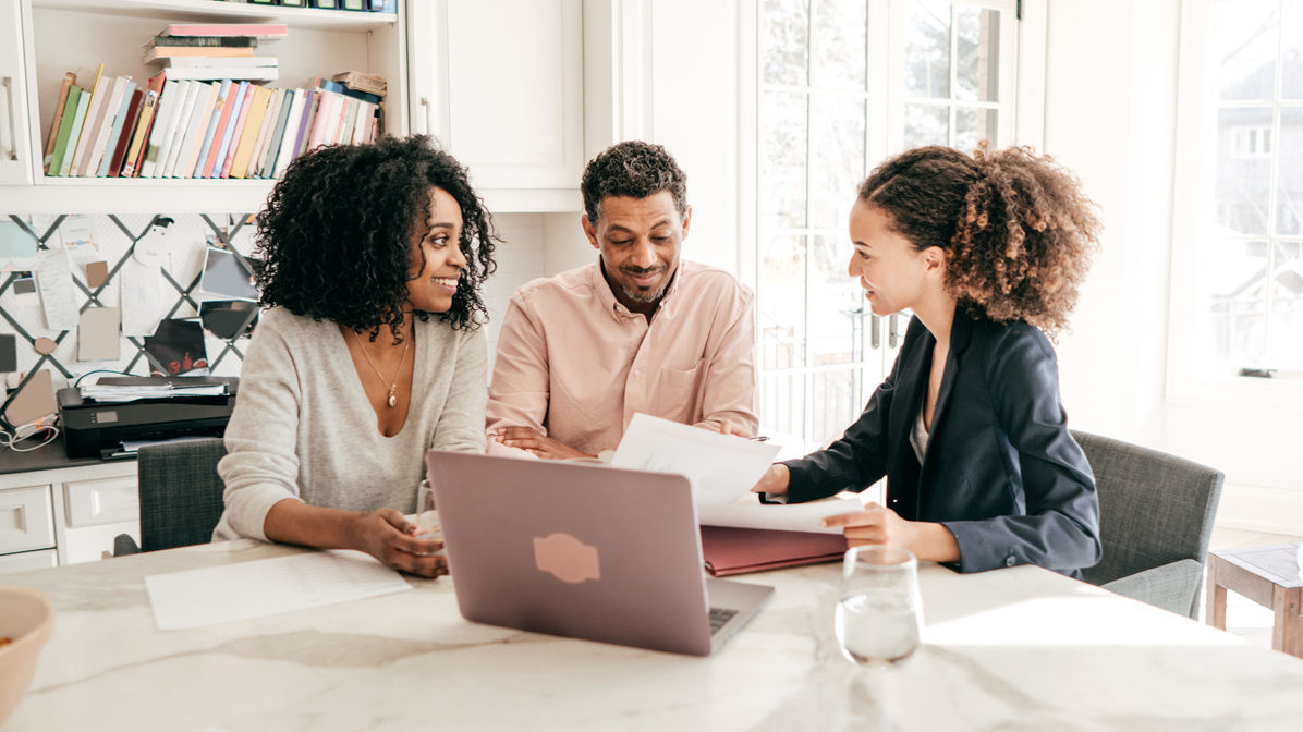 A male and female couple sit at a table with another woman showing them documents.