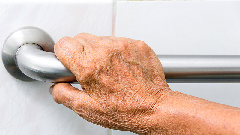 The hand of an elderly person gripping a metal rail on a white tiled wall.