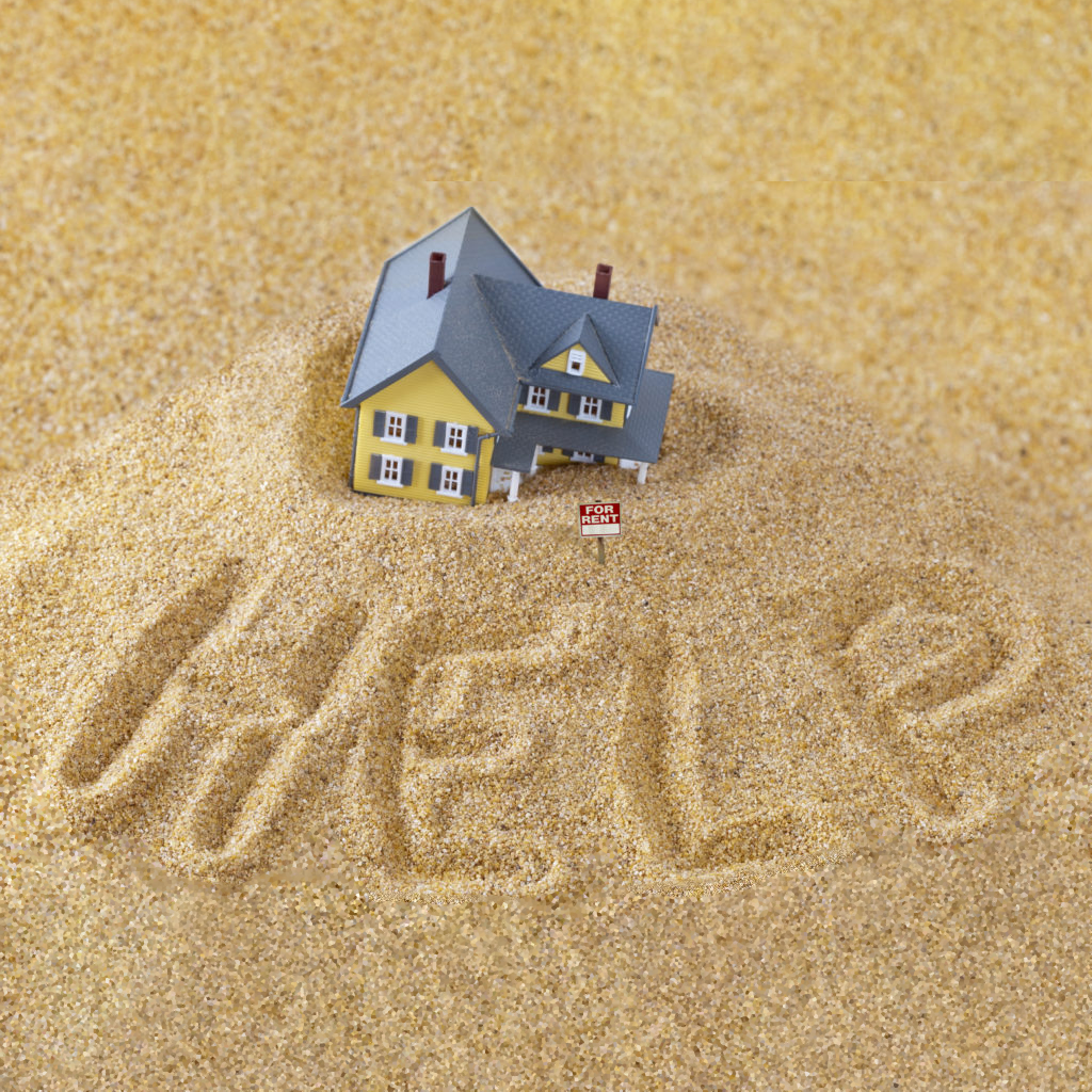 A house sinking in a pile of sand with the word help spelled out in front of it.