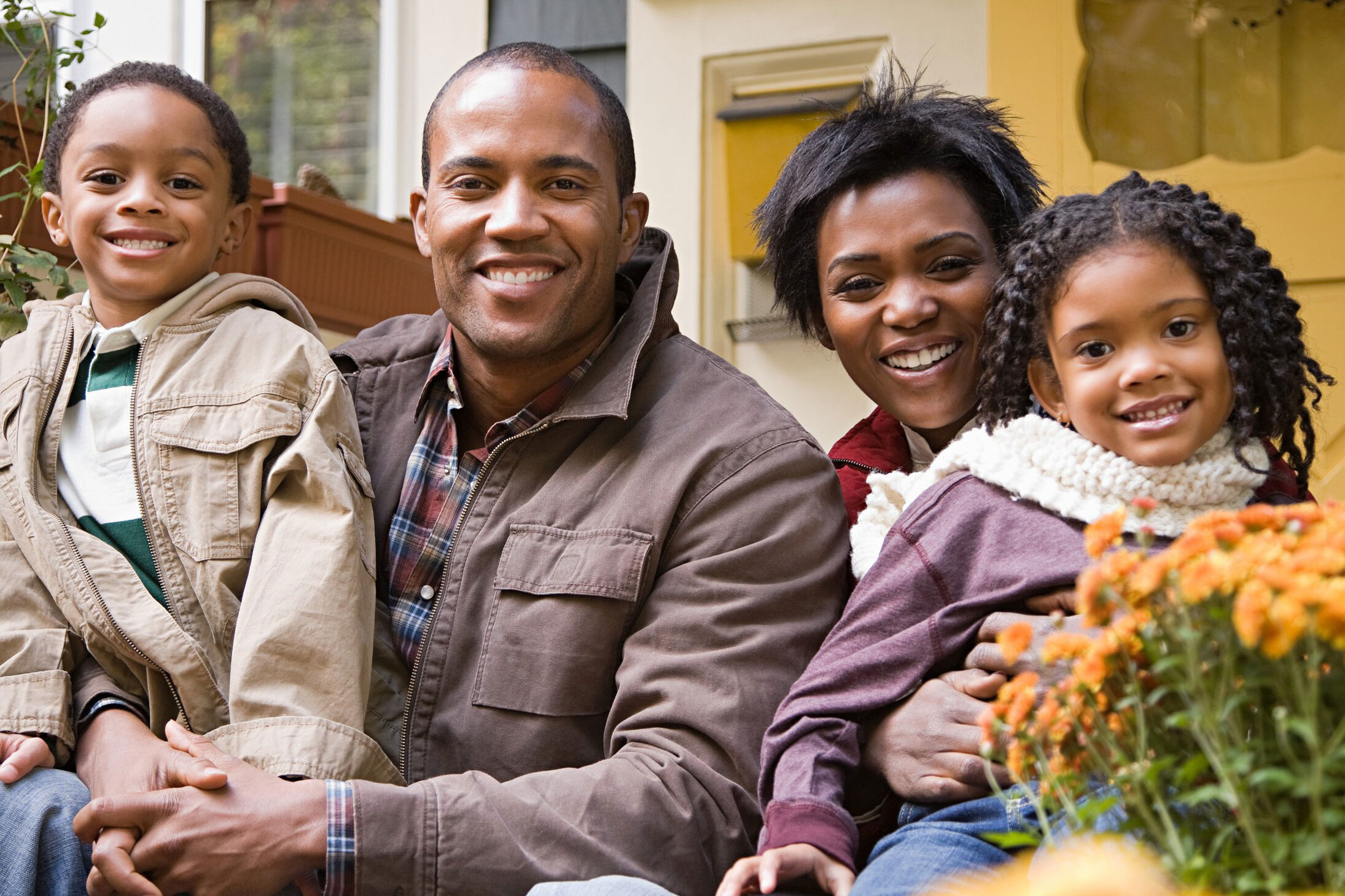 A Black father and mother holding their young son and daughter while smiling as they sit down in front of a home.