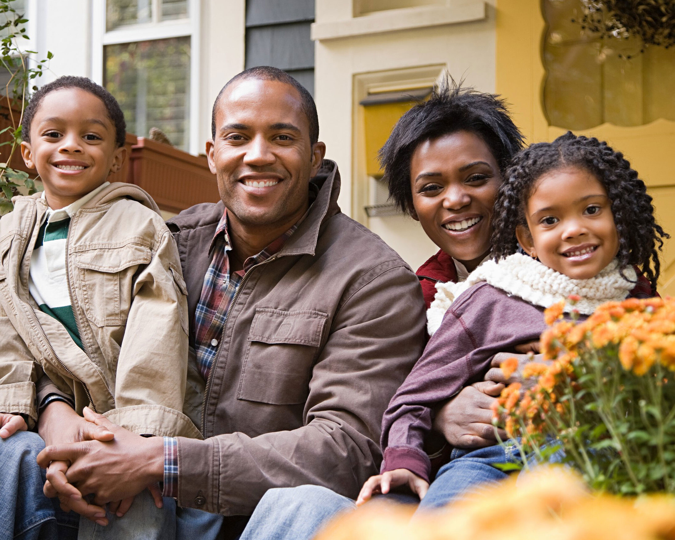 A Black father and mother holding their young son and daughter while smiling as they sit down in front of a home.