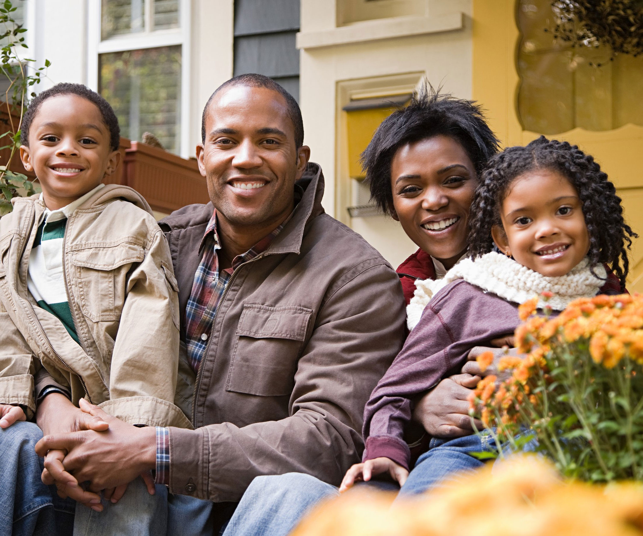 A Black father and mother holding their young son and daughter while smiling as they sit down in front of a home.