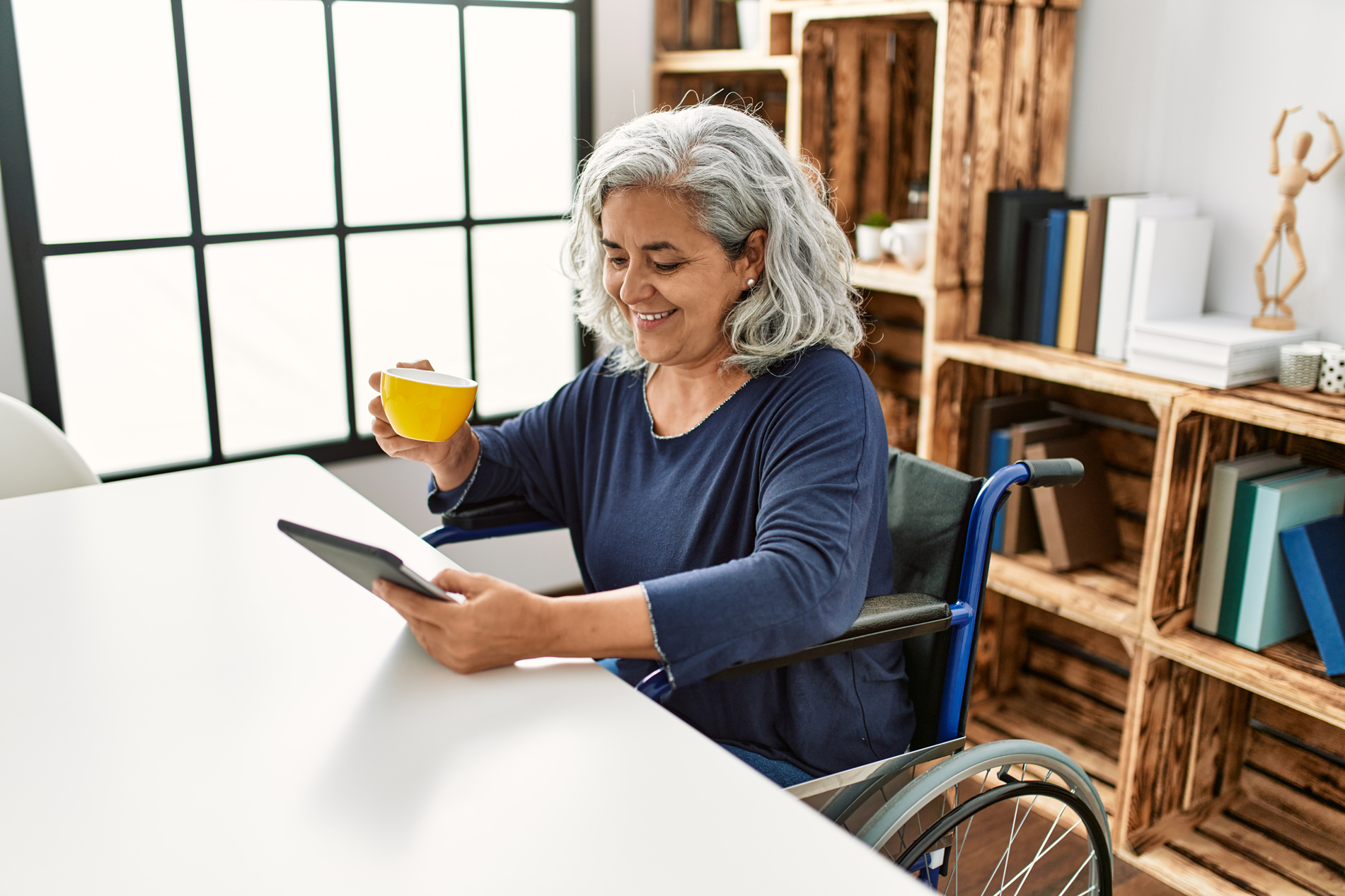 Retired age woman sitting in a wheelchair sipping coffee and reading a screen device